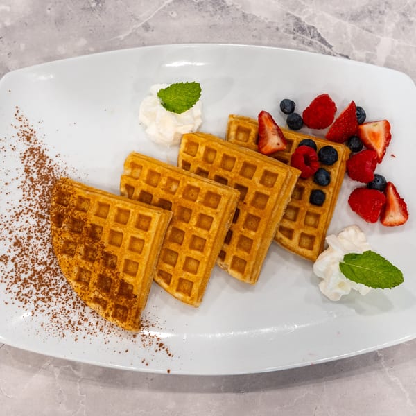 Golden waffles topped with strawberries, raspberries, blueberries, whipped cream, mint, and cinnamon on a white plate.