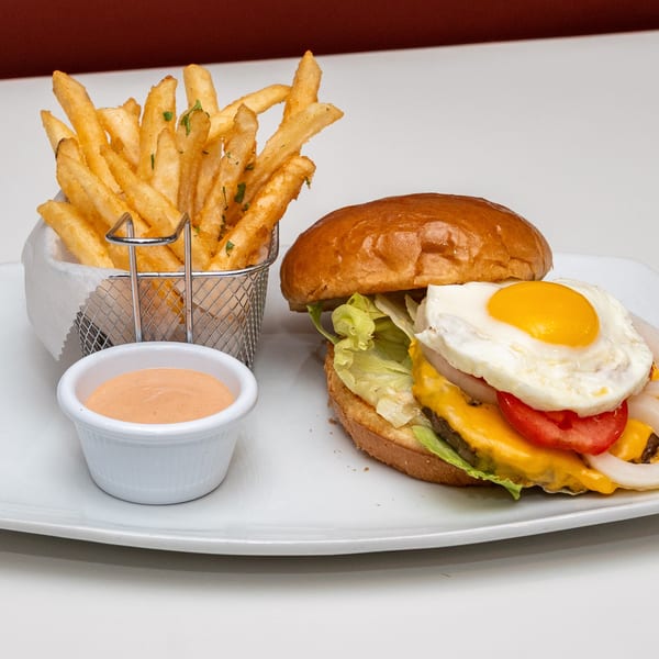 Cheeseburger topped with a fried egg, lettuce, tomato, and onion, served with fries and dipping sauce on a white plate.