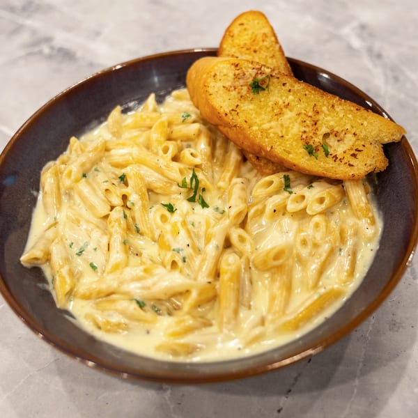Creamy penne pasta garnished with parsley and served with two slices of garlic bread on a dark plate.