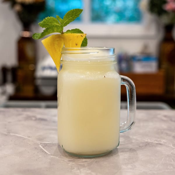 Pale yellow drink in a glass mason jar garnished with pineapple slices and fresh mint on a restaurant table.