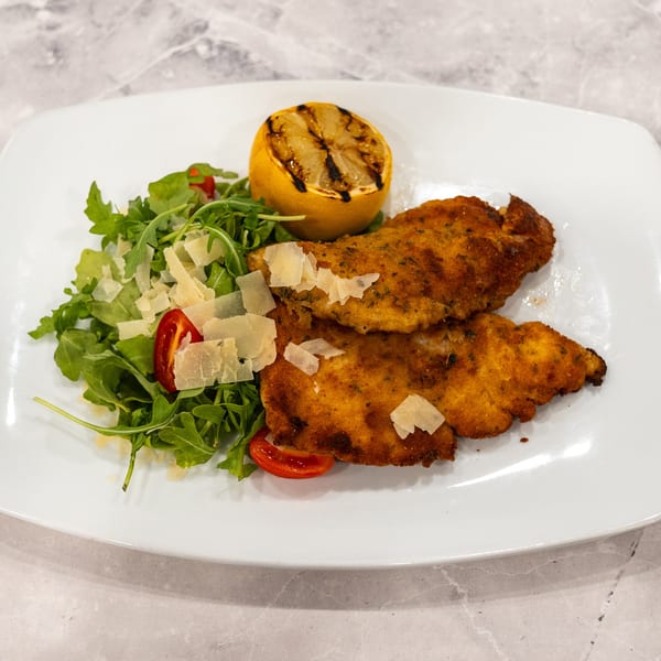 Breaded chicken cutlets with arugula, tomatoes, shaved Parmesan, and grilled lemon on a white plate.